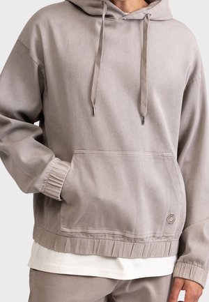Personne portant un sweat à capuche beige avec poche avant, cordons et poignets côtelés, associé à un pantalon assorti et une chemise blanche en dessous.