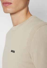 Beige pletený svetr s kulatým výstřihem, se žebrovanými detaily na rukávech a malým černým logem "BOSS" na hrudi.