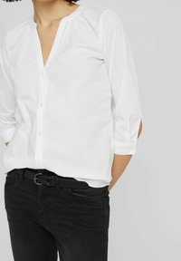 Chemise blanche à col avec manches trois-quarts et patte de boutonnage, associée à un jean noir et une ceinture noire. Tissu doux et lisse.