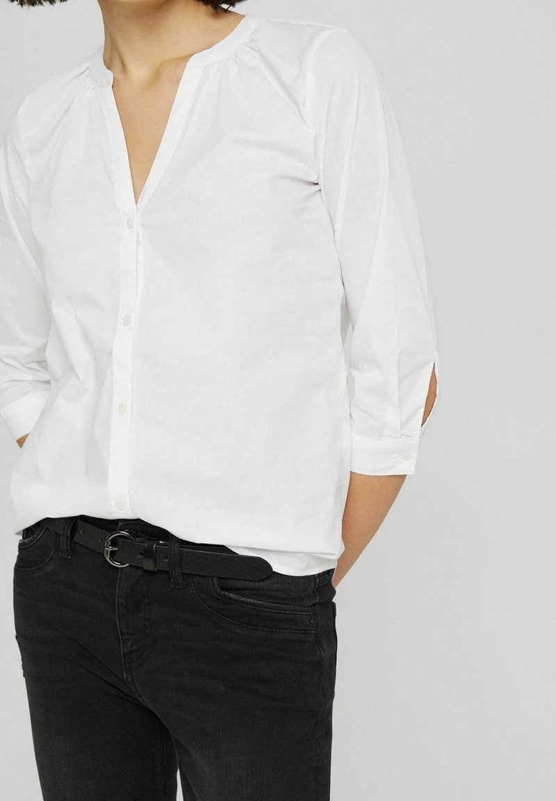 Chemise blanche à col avec manches trois-quarts et patte de boutonnage, associée à un jean noir et une ceinture noire. Tissu doux et lisse.