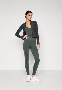 Sort lynlås sportsjakke over en tætsiddende grøn bodysuit og matchende leggings, alle med en subtil glans, parret med hvide sneakers.