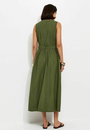 Mujer de cabello oscuro, vistiendo un vestido midi sin mangas verde oliva con cinturón y sandalias negras, de pie contra un fondo blanco y de espaldas.