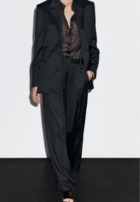 Czarny dopasowany garnitur z wyraźną marynarką i luźnymi spodniami, w połączeniu z ciemną półprzezroczystą koszulą i czarnymi sandałami na paski.