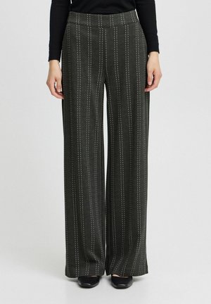 Femme portant un pantalon large gris foncé à rayures verticales blanches, associé à des chaussures noires et un haut noir à manches longues.