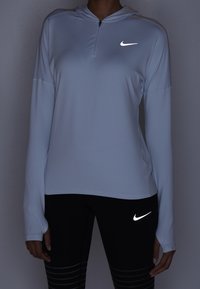 Felpa con zip a maniche lunghe di colore blu chiaro, con logo Nike riflettente sul petto, abbinata a leggings neri con logo riflettente e righe orizzontali.