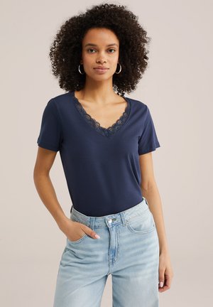 Ženska s kodrastimi lasmi, oblečena v mornarsko modro majico z V-izrezom in čipko ter svetlo modre kavbojke z visokim pasom, stoji z eno roko v žepu.