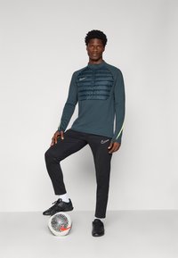 Nike idrettsantrekk bestående av en mørkegrønn langermet topp med en quiltet brystpanel, kombinert med svarte tapered bukser. En fotball ligger under.