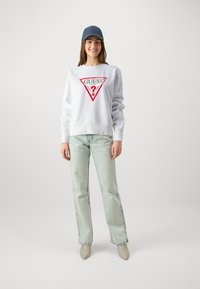Sweatshirt blanc avec logo triangulaire rouge, jean bleu clair et chaussures beige à bout pointu. Le mannequin porte une casquette de baseball noire. Fond neutre.