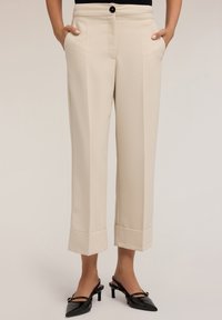 Pantaloni beige su misura con fronte piatto, tasche laterali e risvolti larghi. Abbinati a scarpe con tacco nere lucide. Tessuto dalla texture liscia.