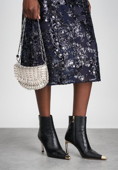 Melnas ādas potītes zābaki ar norādītu pirkstu un zelta akcentiem, kombinēti ar sekvīnēm rotātu jūras zila svārku un mazu krēmbagāžu ar metalizētiem akcentiem.