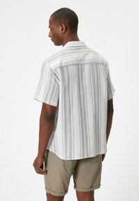 Camisa de manga corta en gris claro con rayas verticales negras, corte relajado, cuello abotonado y textura de tela suave. Usada con pantalones cortos.