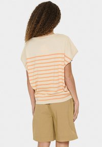 Lichtgewicht kort mouw top in crème met oranje horizontale strepen, met een relaxte pasvorm. Gecombineerd met beige shorts.