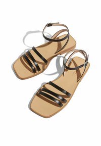 Sandalias planas negras con diseño de tres correas y hebilla en el tobillo, con punta cuadrada y suela beige. Textura de cuero suave.