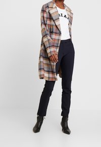 Manteau à carreaux beige, marron et marine ; t-shirt blanc avec texte noir ; pantalon marine ; bottes noires à talon, à bout pointu et finition brillante.