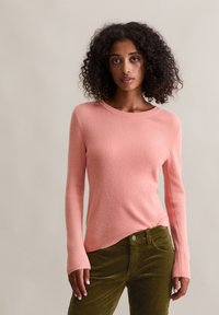 Frau mit lockigem Haar, die einen pinkfarbenen, gerippten Pullover mit langen Ärmeln und olivgrüne Cordhosen trägt, steht vor einem einfarbigen Hintergrund.