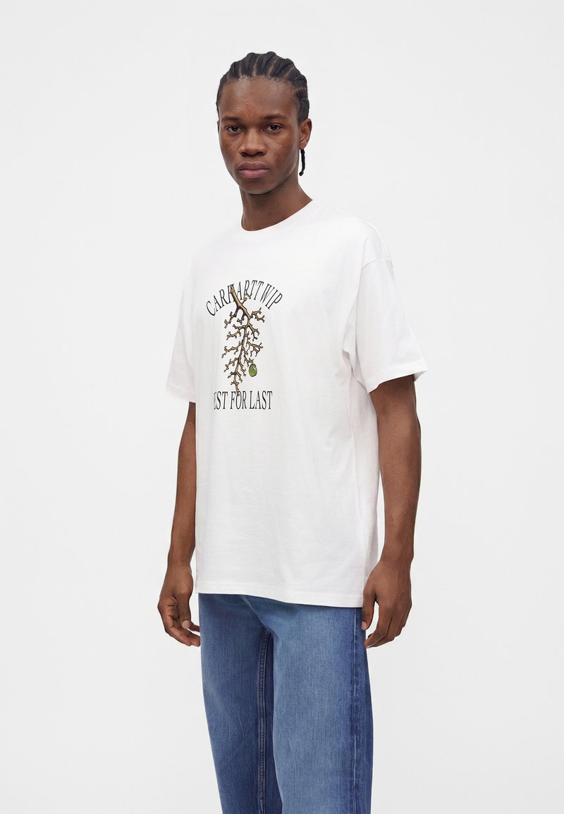 Junger Mann mit geflochtenem Haar, der ein weißes T-Shirt mit Grafik und blaue Jeans trägt, steht vor einem schlichten weißen Hintergrund.