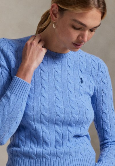 Woman adjusting neckline of light blue cable knit sweater with long sleeves and small embroidered logo on chest.