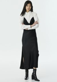 Vestido de satén negro con un escote profundo y una abertura lateral, llevado sobre una camisa blanca con cuello, combinado con botas de tacón brillantes negras.