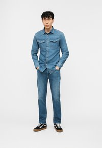 Νέος άνδρας που στέκεται με τα χέρια στις τσέπες, φορώντας μπλε τζιν πουκάμισο, μπλε τζιν παντελόνι και μαύρα αθλητικά παπούτσια, μπροστά σε μια απλή λευκή επιφάνεια φόντου.