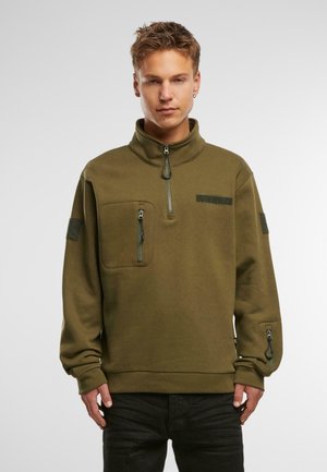 Olivengrøn fleecepullover med høj krave, frontlomme med lynlås og lynlåsdetaljer på ærmerne. Har velcro-detaljer på skuldrene.