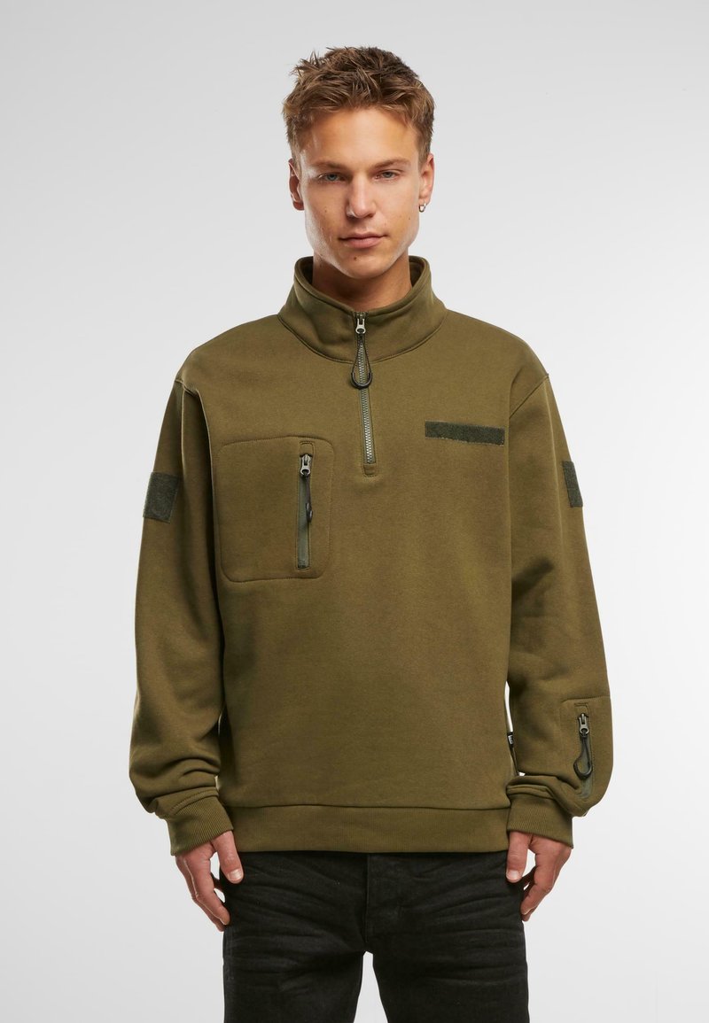Pullover in pile verde oliva con colletto alto, tasca frontale con zip e dettaglio con zip sulle maniche. Presenta accenti in velcro sulle spalle.
