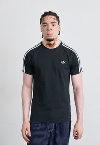 Zwarte katoenen T-shirt met witte drie-strepen detail op de schouders en klein logo op de borst. Getailleerd ontwerp; gedragen met gestreepte broek.