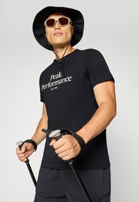 Sort t-shirt med hvid tekst "Peak Performance Est. 1986," parret med en bredskygget sort hat og vandrestave med grå greb.
