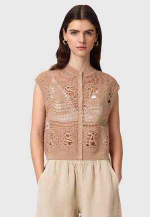 Kvinde med langt brunt hår iført beige hæklede, ærmeløse knap-op top med bogstavmønstre og lysebeige løse bukser, hænderne i lommerne.