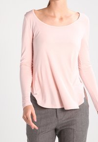 Helles rosa Langarmshirt mit weitem Halsauschnitt und fließendem Saum, getragen über grauen Hosen. Weicher Stoff mit minimalen Nahtdetails.