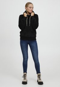 Black, textured hooded sweatshirt with a high collar, drawstrings, and black cuffs, paired with fitted blue jeans and beige hiking boots.