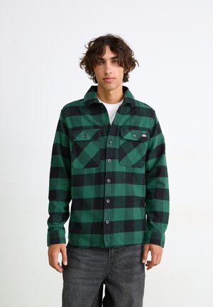 Camisa de franela a cuadros verdes y negros con cuello clásico, dos bolsillos frontales y cierre de botones, llevada sobre una camisa blanca.