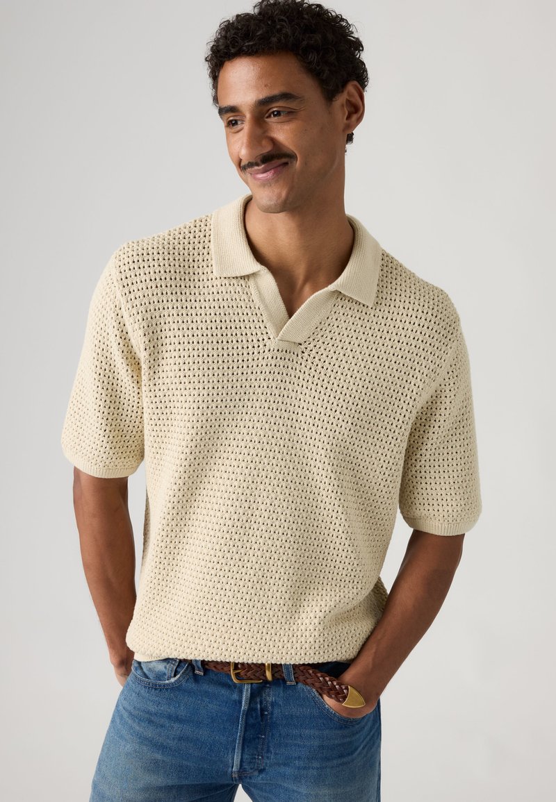 Hombre con cabello rizado y bigote, vestido con una camisa tipo polo de punto beige de manga corta y vaqueros azules, de pie con las manos en los bolsillos, sonriendo.