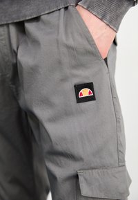 Un pantalon cargo gris présente une poche à rabat avec un logo multicolore brodé. Le matériau semble léger avec une texture lisse.
