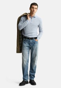 Mann in hellblauem Viertelreißverschlusspullover, ausgebleichten blauen Jeans, schwarzem Gürtel, schwarzen Stiefeln, der einen beigen Mantel mit kariertem Futter über der Schulter trägt.