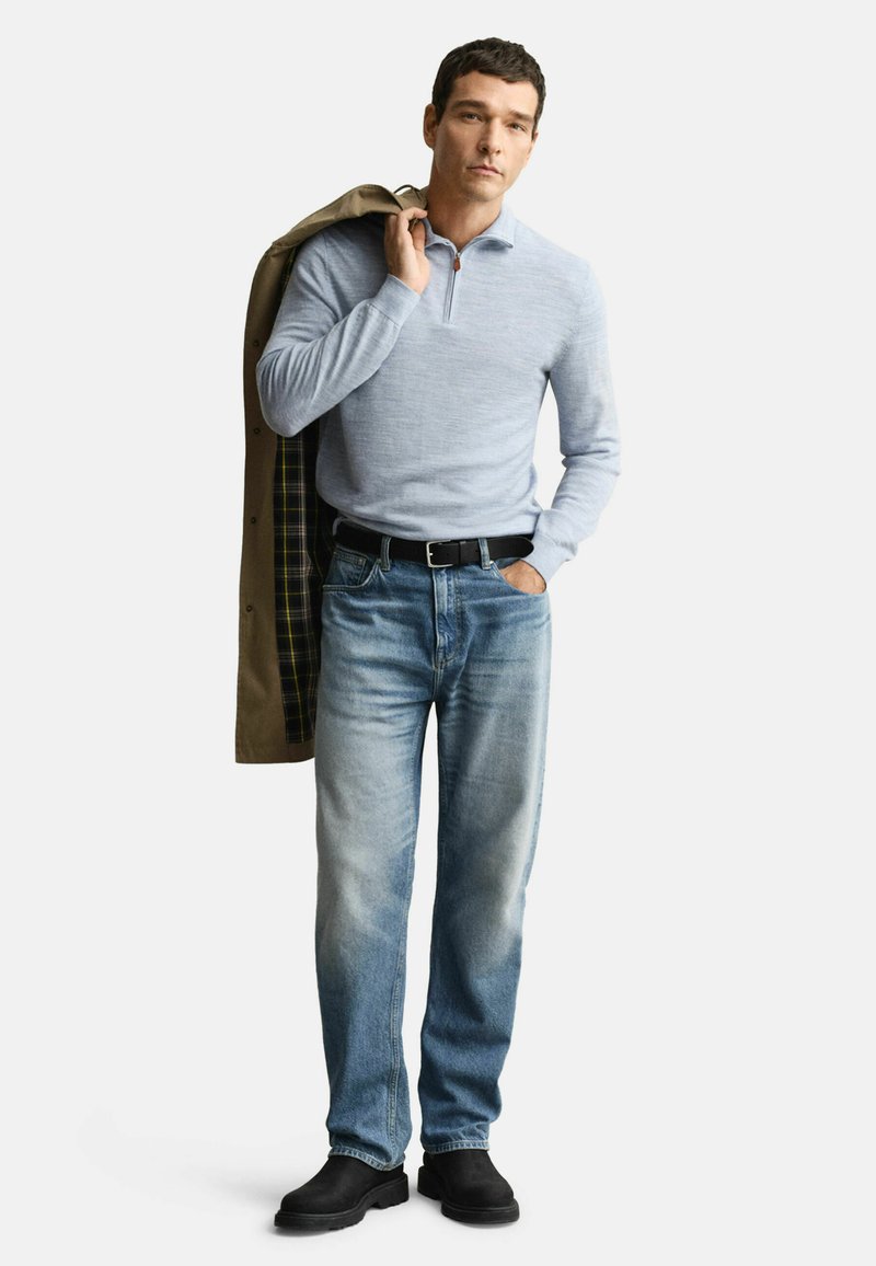 Mann in hellblauem Viertelreißverschlusspullover, ausgebleichten blauen Jeans, schwarzem Gürtel, schwarzen Stiefeln, der einen beigen Mantel mit kariertem Futter über der Schulter trägt.