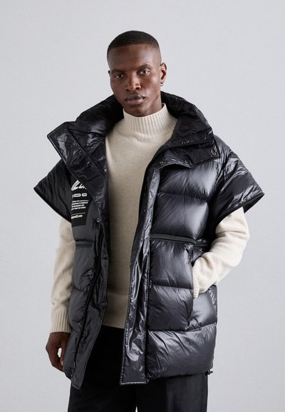 Gilet rembourré noir au design oversize, finition brillante, poches latérales et capuche. Superposé sur un pull en tricot beige.