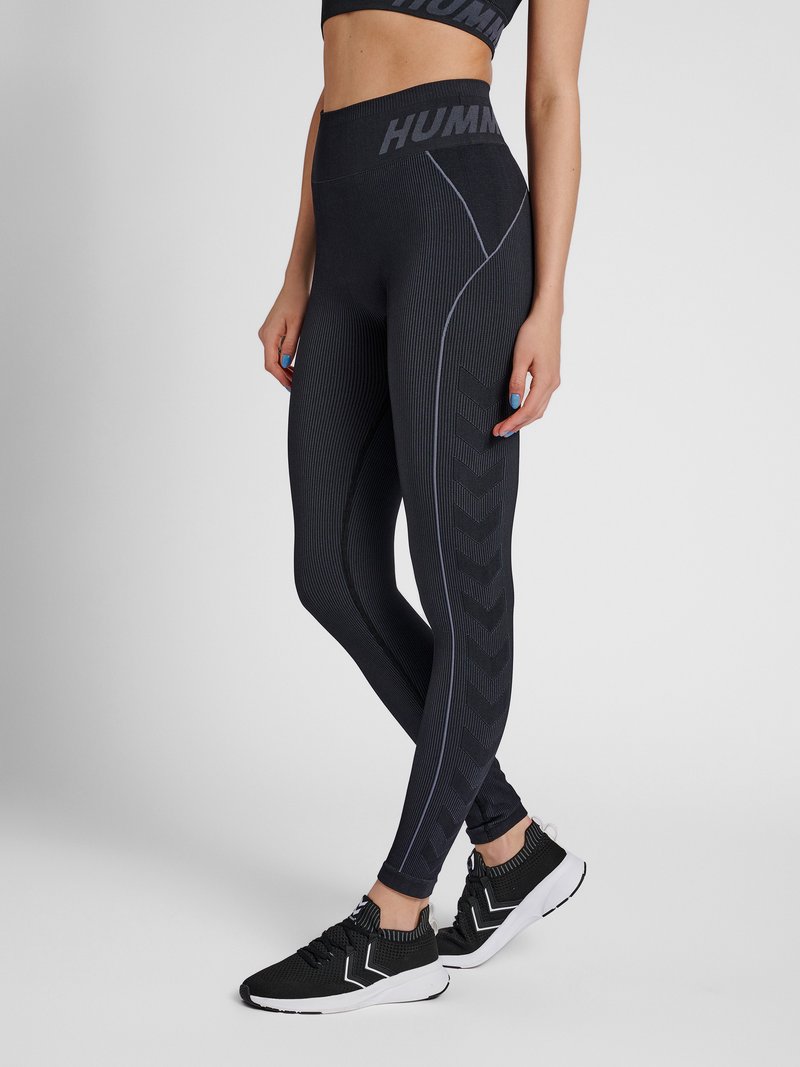 Kvinde iført sorte leggings med høj talje med mærketalje og chevronmønster, kombineret med sorte og hvide sportssko.