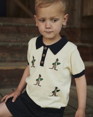 Ung gutt iført en beige polotrøye med svart krage og kenguru surferbroderi, sittende på tretrinn, ser mot siden.