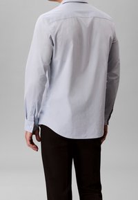 Homme portant une chemise bleu clair à manches longues et un pantalon noir, vu de dos sur un fond gris uni.