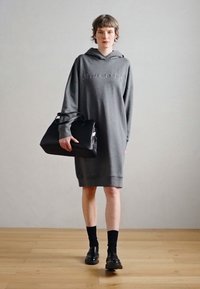 Šedé nadměrné mikinové šaty s reliéfním logem, nošené s černými ponožkami a loafers a držící černou strukturovanou kabelku.