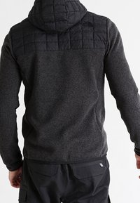 Mies, joka on pukeutunut tummanharmaaseen fleece-huppariin, jossa on tikkaukset mustassa yläselässä ja hupussa, yhdistettynä mustiin cargo-housuihin, seisoo selkä kameraa kohti.