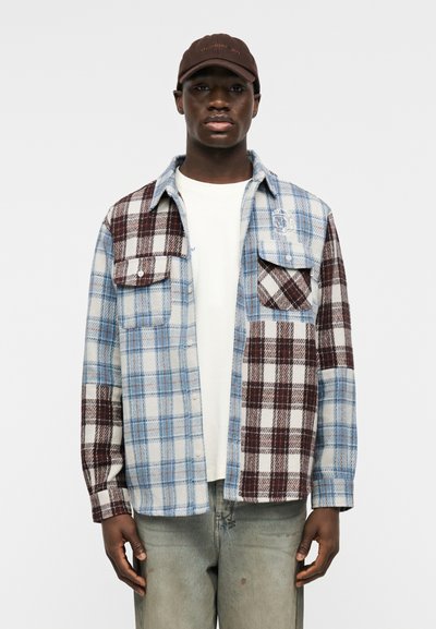 Černoch nosící hnědou čepici, bílé tričko a kostkovanou košili v modro-hnědých tónech s dvěma kapsami na hrudi.