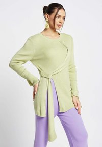 Maglione a coste con maniche lunghe color verde chiaro, caratterizzato da un design drappeggiato unico e un dettaglio annodato, abbinato a pantaloni in lavanda testurizzati.