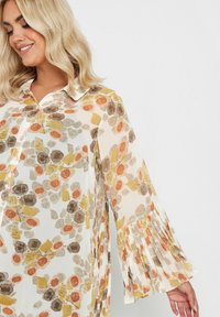 Floral blouse in lightweight, sheer fabric featuring orange, brown, and yellow print. Button-down front and pleated bell sleeves.