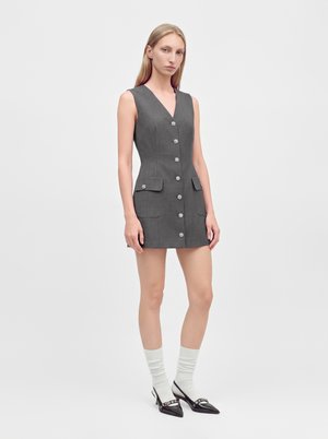 Femme portant une mini-robe grise sans manches boutonnée à l'avant avec des poches, des chaussettes blanches et des chaussures noires à bout pointu, debout sur un fond uni.