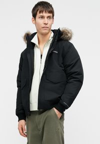 Μαύρο φούτερ μπουφάν με αποσπώμενη κουκούλα επενδεδυμένη με γούνα, δύο μπροστινές τσέπες με καπάκι, ριχτές μανσέτες και φερμουάρ. Φοριέται πάνω από μια λευκή φόρμα.