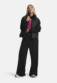 A woman stands confidently, wearing a black puffer vest over a pink top and black pants, with silver sneakers, against a white background.