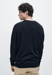 Černý pletený svetr s kulatým výstřihem a dlouhými rukávy, s volnějším střihem a hladkou texturou. Nošený přes světle barevné kalhoty.