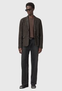 Uomo con occhiali da sole neri, giacca marrone scuro, maglione marrone, cintura nera, jeans grigio scuro e stivali neri che si trova davanti a uno sfondo grigio.