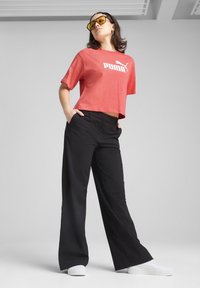 Rød cropped t-shirt med hvidt Puma-logo, parret med løse sorte bukser og hvide sneakers. Model iført gule solbriller.